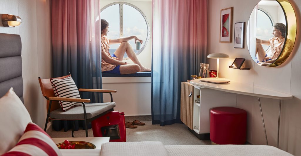 Un chico se relaja y mira por la ventana en su camarote del crucero Resilient Lady de Virgin Voyages
