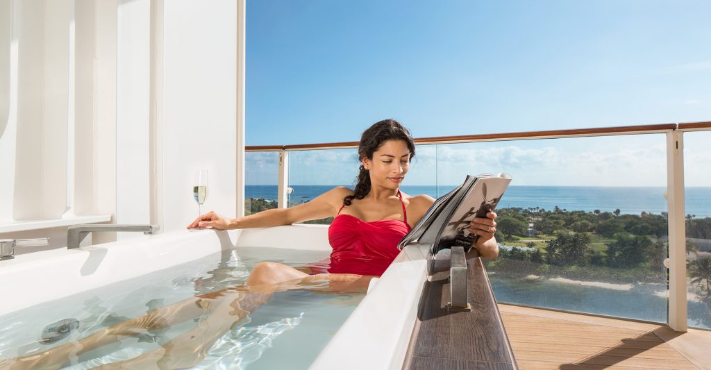 Una mujer se relaja con una revista en el jacuzzi de su suite en un crucero Celebrity Imagen de una mujer leyendo una revista, desconectando de su móvil, en el jacuzzi de su suite en un crucero Celebrity