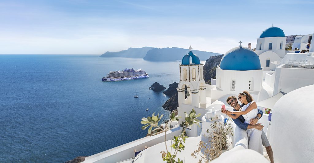 Reservar con agencia especializada en cruceros: Una pareja disfruta de su escala en Santorini con un barco de la naviera NCL detrás