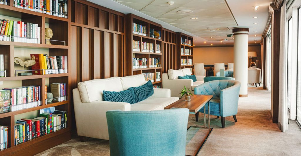 Sala de lectura del crucero Azamara Onward con biblioteca, sofás y ventanales al mar