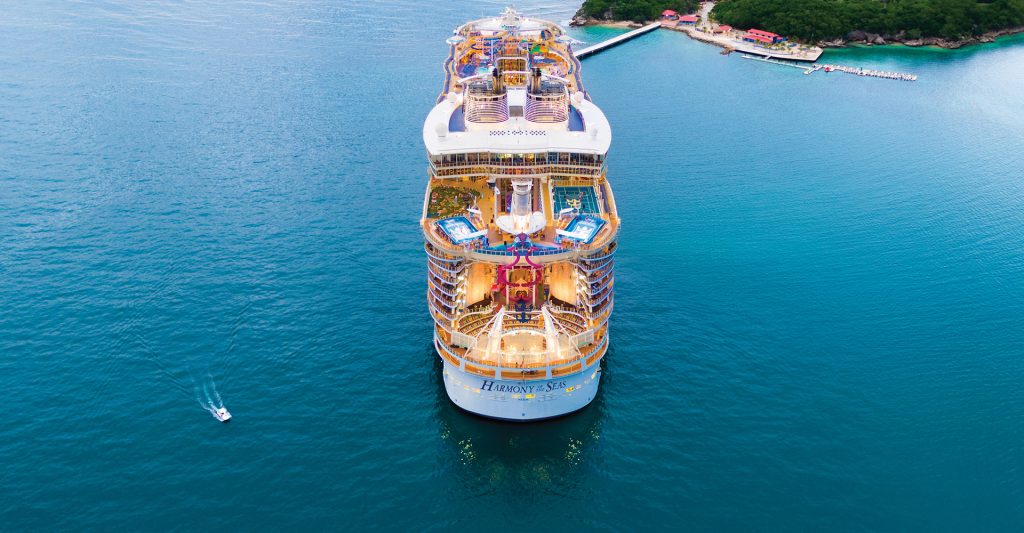 Vista aérea de la popa de un gigantesco crucero de la clase Icon de Royal Caribbean amarrado junto a un exuberante muelle tropical rodeado de aguas turquesas.