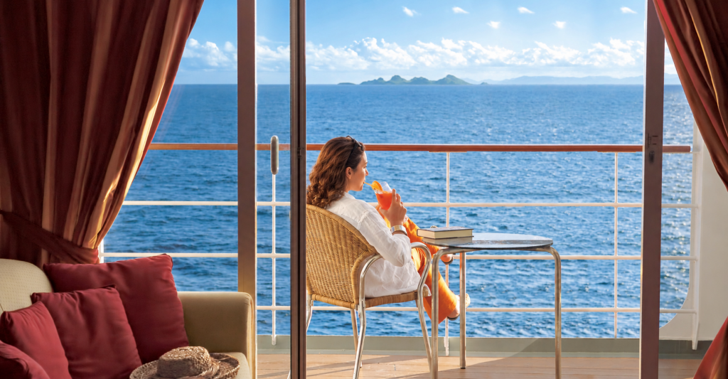 camarote-con-balcon-crucero-vistas-al-mar Mujer relajándose en la terraza de un camarote de crucero tomando un refresco con vistas al mar