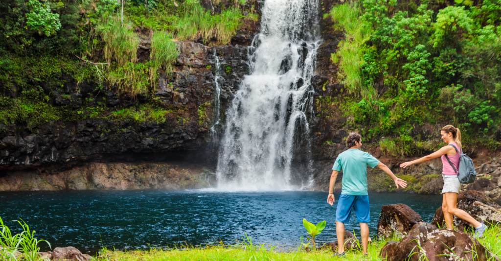 Ruta de senderismo en pareja hacia una cascada natural Pareja haciendo senderismo frente a una cascada natural y una poza de agua cristalina rodeada de vegetación.